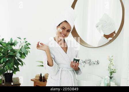 A smiling woman in a bathrobe and a towel on her head applies moisturizer from a jar to her face in the bathroom. The concept of skin care, moisturizing and cleansing the face and neck. Stock Photo