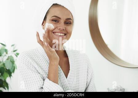 A beautiful young woman with vitiligo on her hands in a bathrobe and with a towel on her head applies moisturizer to her face. The concept of skin care, moisturizing and cleansing the face and neck. Stock Photo