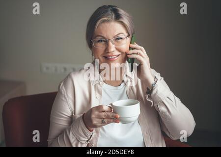 Caucasian senior lady with glasses holding a cup of tea is smiling while talking on phone at home Stock Photo