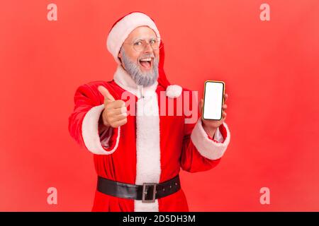 Happy satisfied man with gray beard in santa claus costume holding smartphone with empty white display and showing thumbs up. Indoor studio shot isola Stock Photo