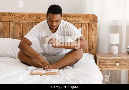 African Man Reading Book Drinking Morning Coffee Sitting In Bedroom Stock Photo