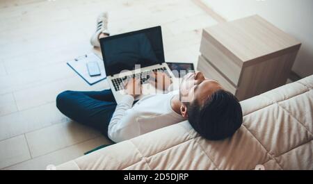 Young caucasian businessman feeling tired after working on the laptop from home sitting on the floor near the bed falling asleep Stock Photo
