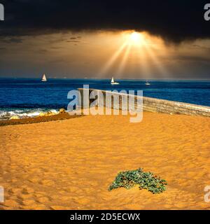 Bright sunlight breaks through clouds over a sandy beach with sailing boats and a seaside pier at dusk, Oleron, Charente-Maritime, France Stock Photo