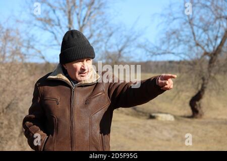 Elderly man in warm clothes points to something in the distance. Rural scene, concept of old age, life in village Stock Photo