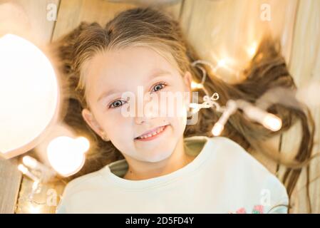 Happy girl lies on the wooden floor in her hair with a bright garland of Christmas lights. Smiling, looking at the camera. Shooting from above, Stock Photo