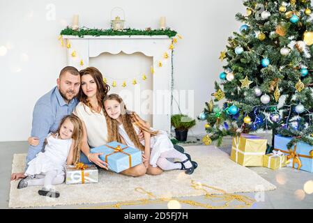 Happy family are holding boxes with gifts in front of a white fireplace with an elegant Christmas tree in garlands of lights. Young father and beautif Stock Photo