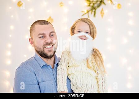 Close-up portrait of happy dad and blonde daughter in front of a Christmas tree with a garland of lights. The daughter put on a santa claus beard. Stock Photo