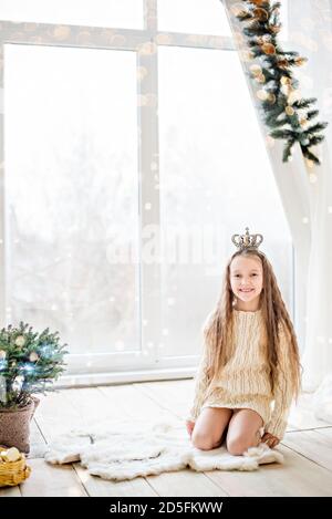 Close-up portrait of a little blonde girl a white knitted sweater dress with a crown on her head. She smiles and looks into the camera. The Snow Queen Stock Photo