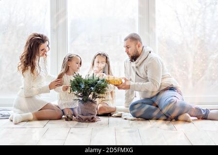 A happy family in white sweaters prepares to meet New Year in a beautiful loft with panoramic windows. Young parents with little daughters decorate a Stock Photo