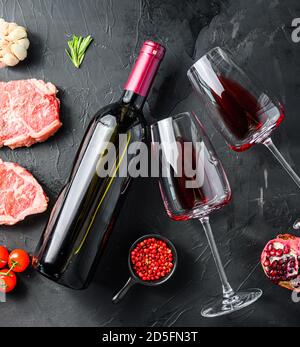 Red wine bottle with two glasses near top blade steaks with herbs seasoning over black textured background top view square. Stock Photo