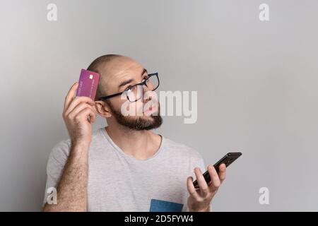 The concept of banking and credit card purchases. A man with glasses holds a Bank card in one hand and a mobile phone in the other. Emotion of thought Stock Photo