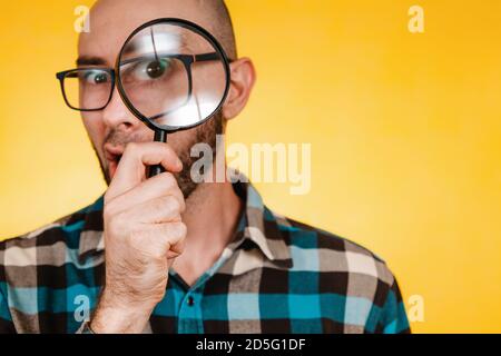 The concept of finding and solving problems. A bald man with a beard and glasses, wearing a blue checked shirt, looks through a magnifying glass. Eye Stock Photo