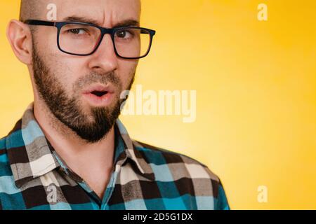 People and emotions. A bearded man with glasses and a blue checked shirt asks something in a dissatisfied voice. Yellow background. Copy space.. Stock Photo