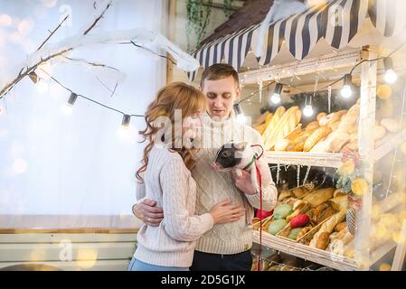 A happy family in white knitted sweaters stands at the bakery, choosing pastries, laughing, holding a mini pig. The showcase is  Christmas tree Stock Photo