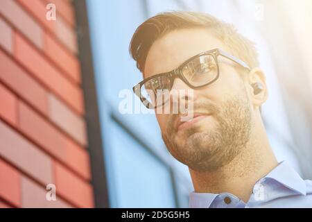 Portrait of a young handsome businessman wearing black wireless earphones listening to music, enjoying favourite song while standing outdoors Stock Photo