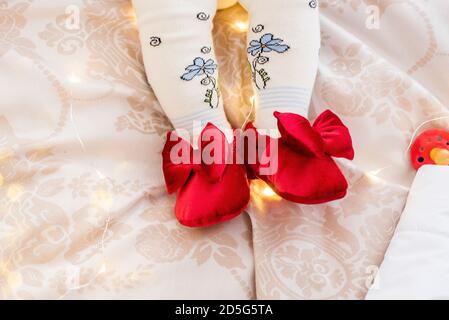 On small legs, the baby is wearing white tights and red velvet booties. Festive shoes for Christmas in garlands of lights. Greeting card, copy space, Stock Photo