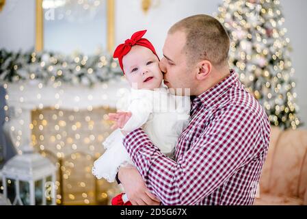 A young man holds a baby in his arms. Father plays with a girl in a white dress, hugs her, kisses against the background of festive Christmas trees Stock Photo