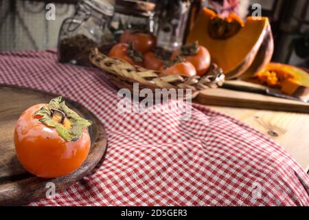 A persimmon resting on a wooden cutting board and a red and white checkered tablecloth, in the background other persimmons and a pumpkin Stock Photo