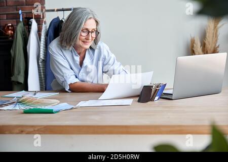 Middle aged stylish woman fashion designer holding sketches in studio office. Stock Photo