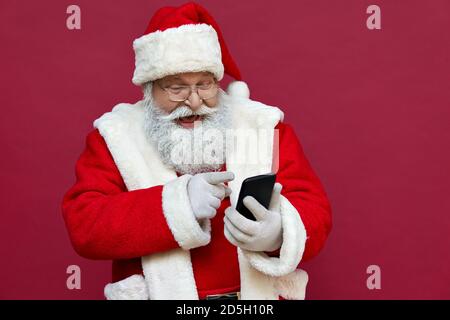 Happy excited Santa Claus using smart phone isolated on red background. Stock Photo