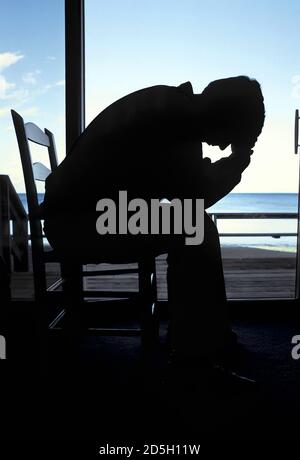 Silhouette of an adult male sitting in a chair with head in hand in deep depression comtemplating suicide Stock Photo