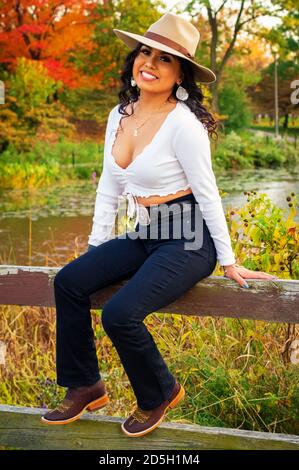 Young Mexican woman is wearing a hat and boots while sitting on a bridge Stock Photo