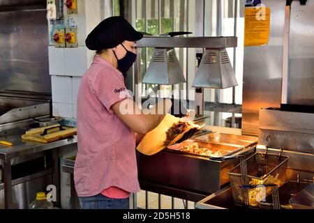 Young woman preparing food (a gyros, kebab) for a customer while wearing a face mask and gloves due to covid-19 precautions Stock Photo