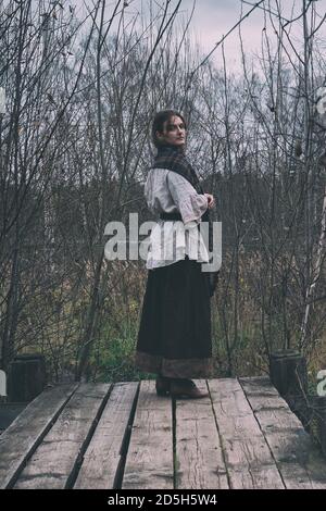 A woman in a headscarf looks from the bridge at the old pond. Girl in a retro dress on a platform in an autumn gloomy landscape Stock Photo