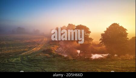 Autumn foggy sunrise. Colorful misty morning dawn. Fall rural scene panorama. Small river with trees in meadow and field, Belarus Stock Photo