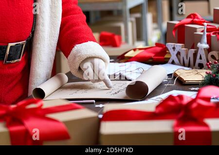 Santa Claus reading wish list standing at table in workshop, close up. Stock Photo