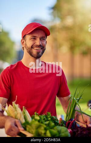 Young happy handsome caucasian male courier in red uniform smiling wide while delivering fresh green grocery. Food delivery concept Stock Photo