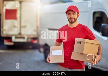 Young caucasian male courier in red uniform smiling at camera while holding small parcels standing on trucks background in yard. Food delivery concept Stock Photo