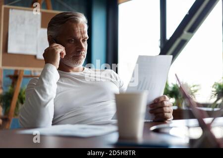Grey-haired mature businessman sitting at table, working on computer, talking on smartphone. Telephone conversations. Stock Photo