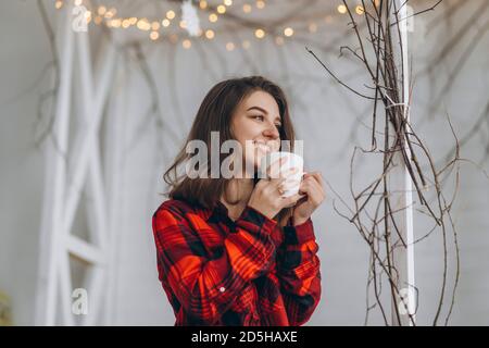 Pretty brunette girl in bed wearing red shirt and warm socks with cup of coffee and christmas lights behind Stock Photo