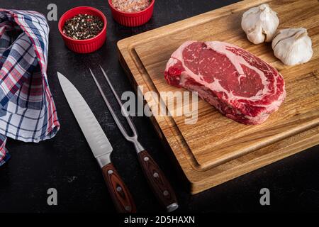 Photograph of raw boneless ribeye steak with sea salt, pepper, garlic on a cutting board with a carving knife and fork set Stock Photo