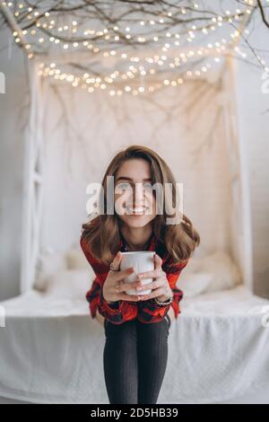 Pretty brunette girl in bed wearing red shirt and warm socks with cup of coffee and christmas lights behind Stock Photo