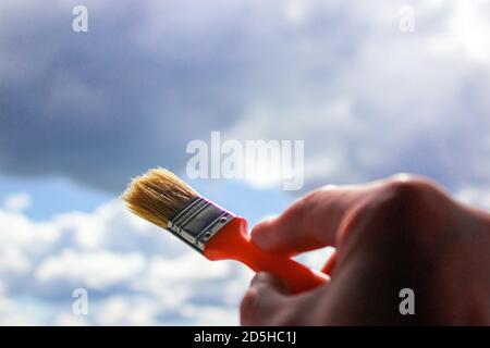Close-up of hand holding paint brush against sky. Clouds painting. Concept. Sunshine and clouds. Stock Photo