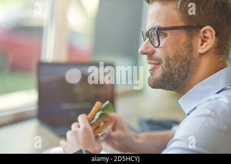 Side view of a young happy businessman sitting at wooden table in cafe and going to eat fresh sandwich, working remotely Stock Photo