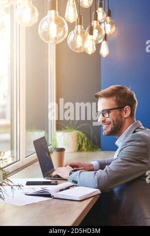 Happy freelancer. Side view of a young cheerful businessman using laptop and smiling while working remotely in cafe Stock Photo