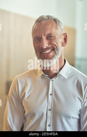 Vertical shot of a happy mature businessman in classic wear looking at camera and smiling while standing in the office Stock Photo