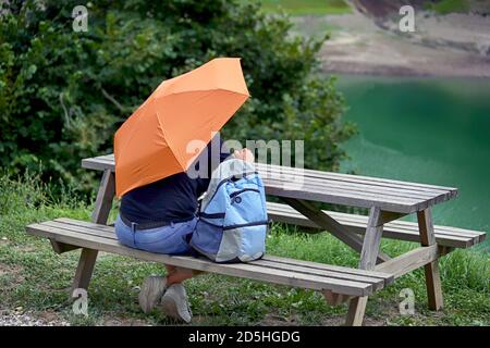 Rear view of a man with an open umbrella and backpack who sitting at a wooden picnic table on a green lawn with trees and a lake in the background. Stock Photo