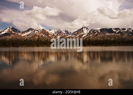 Jagged snowy mountain peaks with snow on the top reflected in a shallow river during early summer. Stock Photo