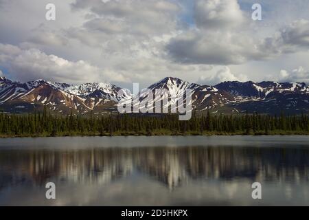 Jagged snowy mountain peaks with snow on the top reflected in shallow river during early summer. Stock Photo