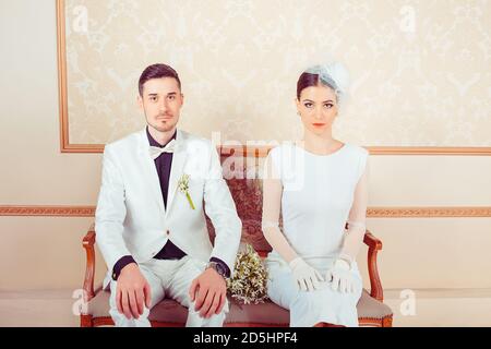 Content elegant man and woman in wedding outfits sitting on sofa in ornamental interior looking at camera Stock Photo