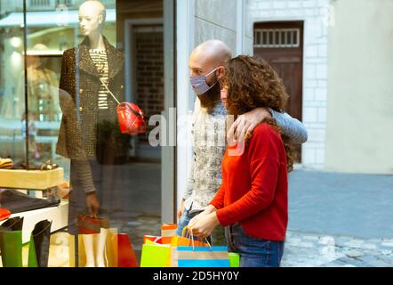 side view of a caucasian couple of a bearded man and a curly woman walking past a shop window and looking in wearing face mask. Stock Photo