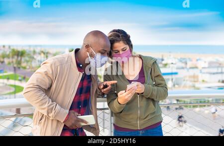 happy interracial couple wearing face mask interacting with smatphones on the seaside in a autumn holidays . concept about the new normality lifestyle Stock Photo