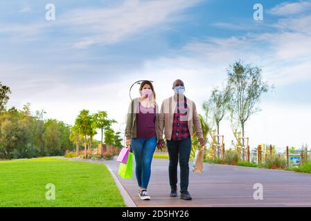 attractive interracial couple wearing face mask holding hands with shopping paper bags whilst walking in a park in autumn.concept about the new normal Stock Photo