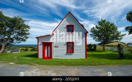Side view of a vintage cottage under blue sky and clouds. The small white wooden building has two double hung windows with red trim. Stock Photo