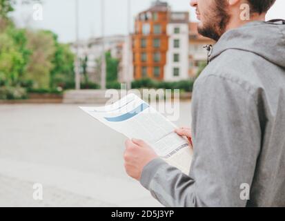 Hands of a man in gray hoodie holding a newspaper looking away to the side thoughtful, daydreaming isolated on city street background Stock Photo