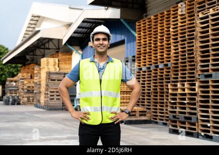 Portrait of young Indian worker working in logistic industry outdoor in front of factory warehouse. Smiling happy man with industrial hard hat looking at camera hands on hip at depot Stock Photo